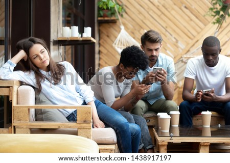 Unhappy millennial girl sit alone aside feel bored at group meeting with friends in café, diverse young people busy using smartphones addicted to gadgets, annoyed female lack communication Royalty-Free Stock Photo #1438917197