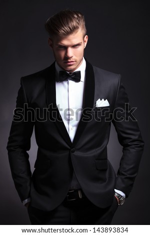 elegant young fashion man in tuxedo is holding both hands in his his pockets and looking at the camera.on black background Royalty-Free Stock Photo #143893834