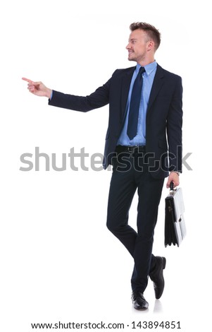 full length picture of a young business man holding a suitcase and pointing and looking to his side. on white background