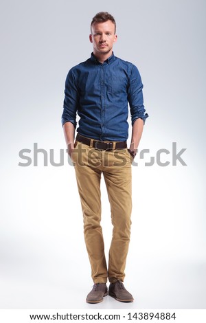 full length picture of a casual young man standing with his hands in his pockets while looking into the camera with a serious expression. on gray background