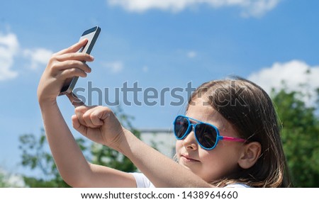 Young brunette girl in summer day enjoying the sun, making selfie portrait. Baby holds a mobile phone in hand and smiles. Little child using smartphone. Close up. Blue sky background with clouds