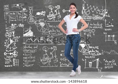 full length picture of a young casual woman standing with her hands on her hips in front of a background full of sketches and charts and smiling for the camera