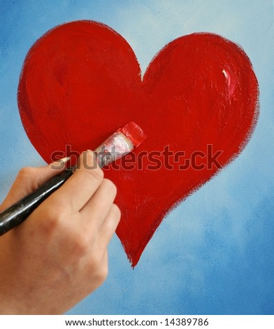 A female hand is painting a red heart on blue background with paintbrush and acrylic color.