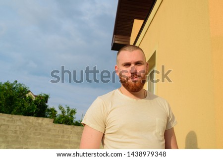 Male with trendy hairdo and beard, looks with serious expression, has thick red beard. Red-bearded man on the street in summer. Male 28-35 years old. The guy against the sky. Royalty-Free Stock Photo #1438979348