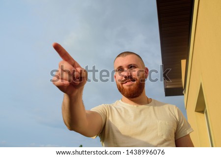 Male with trendy hairdo and beard, looks with serious expression, has thick red beard. Red-bearded man on the street in summer.The guy against the sky. Man points finger. Male 28-35 years old. Royalty-Free Stock Photo #1438996076