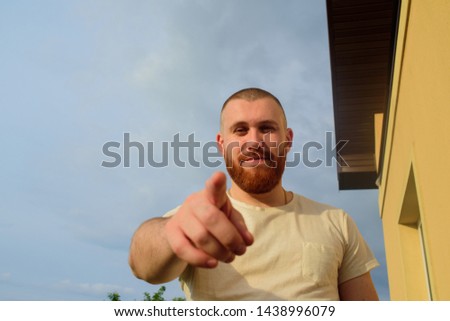 Male with trendy hairdo and beard, looks with serious expression, has thick red beard. Red-bearded man on the street in summer.The guy against the sky. Man points finger. Male 28-35 years old. Royalty-Free Stock Photo #1438996079