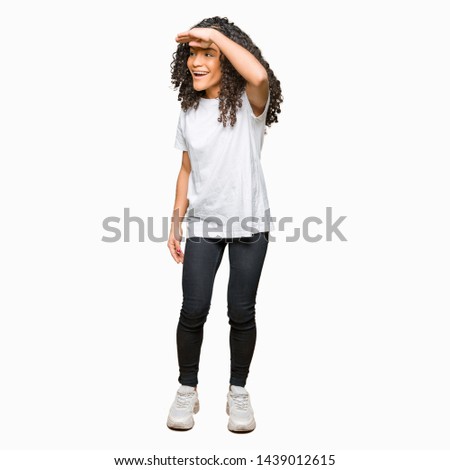 Young beautiful woman with curly hair wearing white t-shirt very happy and smiling looking far away with hand over head. Searching concept.