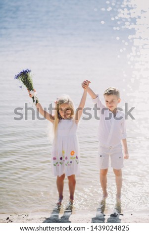 Little boy and girl in a white dress with a hat with a beautiful bouquet of flowers walking along a sandy beach