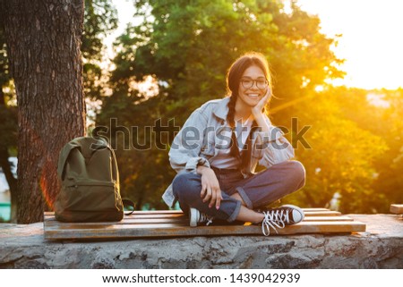 Photo of a pleased happy cute young student girl wearing eyeglasses sitting on bench outdoors in nature park with beautiful sunlight.