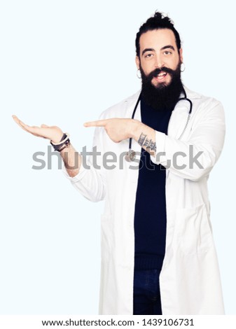 Doctor with long hair wearing medical coat and stethoscope amazed and smiling to the camera while presenting with hand and pointing with finger.