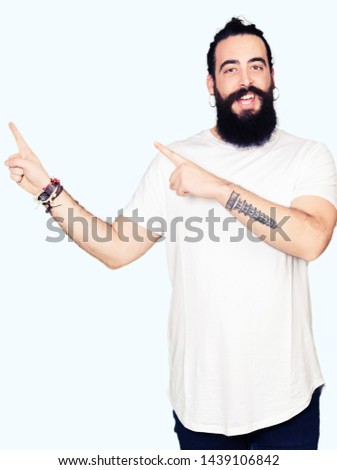 Young hipster man with long hair and beard wearing casual white t-shirt smiling and looking at the camera pointing with two hands and fingers to the side.