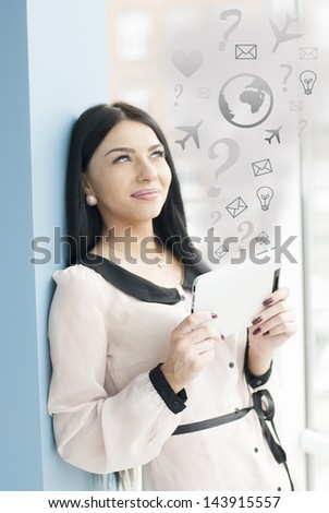 Portrait of a happy smiling young business woman using tablet PC while standing relaxed near window at her office with ideas sign around her