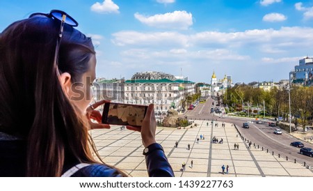 A girl taking a picture of a square with her phone, from a tower above the square. Lots of people walking by on the square. In the back there is St Michael's Golden-Domed Cathedral, Kiev.