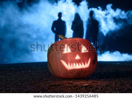 orange pumpkin with slots in the form of eye sockets and mouth on the background of silhouettes of people in thick fog and bright light