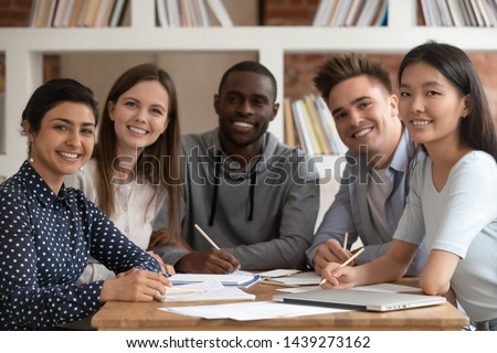 Group picture of happy multiethnic young people sit at shared desk look at camera studying together, multicultural excited students or groupmates smiling posing for photo working in library