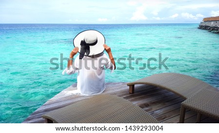 Young Asian woman in white clothes and hat sitting on a wooden balcony over the turquoise water sea for summer holiday vacations concept.