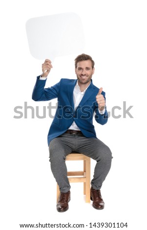 Attractive casual guy holding a speech bubble above his head and giving a thumbs up while wearing an elegant suit and sitting on white studio background