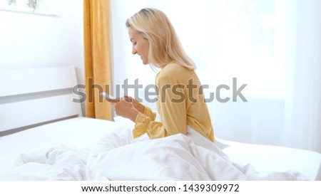 Beautiful blonde woman wearing pajama using phone in her bedroom. 