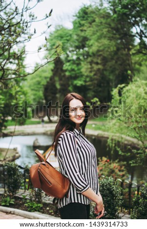 A girl in a striped black and white shirt holding a brown bag and looking at the camera. Great green park view behind her head.