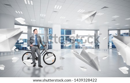 Young man wearing business suit and tie with bike standing at comfortable coworking space with flying paper planes. Happy businessman with bicycle at modern office interior with big windows.