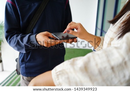 Woman giving handwritten signature with a smartphone or digital signing for goods received from delivery man in front of the glass door with green floor. (selective focus, blurred space for text)