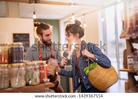 Beautiful young woman shopping in a bulk food store. The seller advises her to buy organic spices in jars. Royalty-Free Stock Photo #1439367554