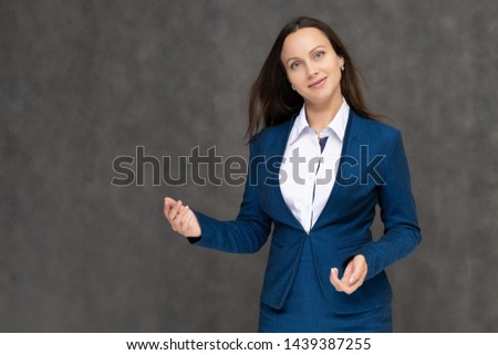 Portrait to the waist a young pretty brunette manager woman of 30 years in a business blue suit with beautiful dark hair. It is standing on a gray background, talking, showing hands, with emotions