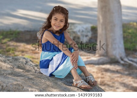 A young girl in a long dress sits on a large rock under a tree in a Florida park. She looks at the camera with a pleasant smile with hand placed on her knees.