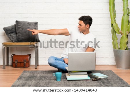 Handsome man sitting on the floor with his laptop pointing finger to the side