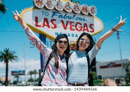 Attractive gorgeous travel girl partners with beaming grins and posing with victory finger sign outside. excited female backpackers on sunny day taking picture face camera with las veags billboard.