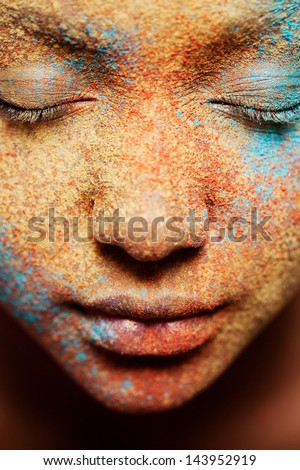 Close up beauty portrait of a young girl face wearing golden yellow, red and blue and make up powder pigment covering her face, detail texture view.