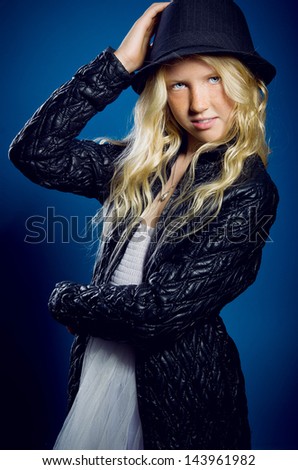Beautiful blonde girl wearing a hat and a black jacket on a blue background. Dreamy girl holds the hand hat
