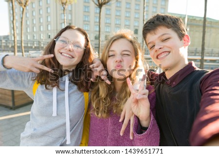Portrait of friends teen boy and two girls smiling, making funny faces, showing victory sign in the street. City background, golden hour