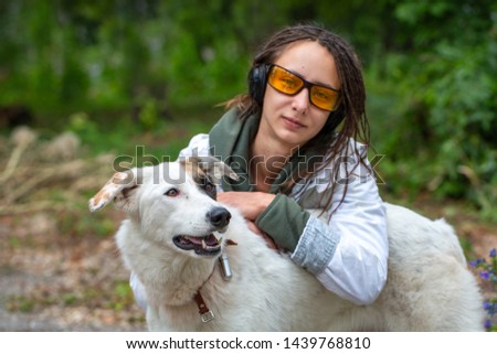 Girl in headphones and glasses hugs a dog. Latino girl of appearance with dreadlocks wearing a white windbreaker. Summer day. The joy and pleasure of communicating with animals.