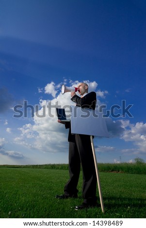 Bald man in a business suit outside in a grassy field holding a computer, a bullhorn and a blank sign, caucasian/white.