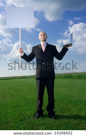 Bold man standing on the grass. He hold blank sign in one hand and laptop in other. Available space for text.