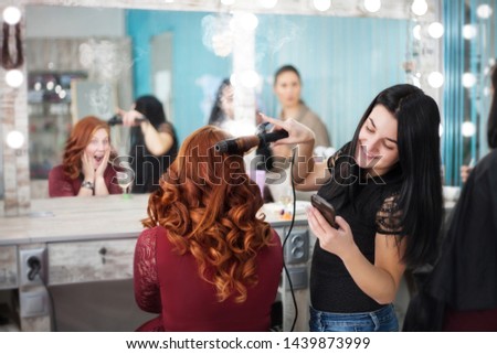 Comic photo in a beauty salon. The hairdresser looks into the phone and the client's hair is on fire. Young women in the beauty salon. Hair and makeup.