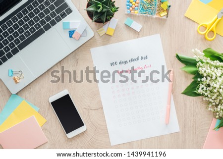top view of 100 days check list with smartphone, laptop and stationery on wooden table