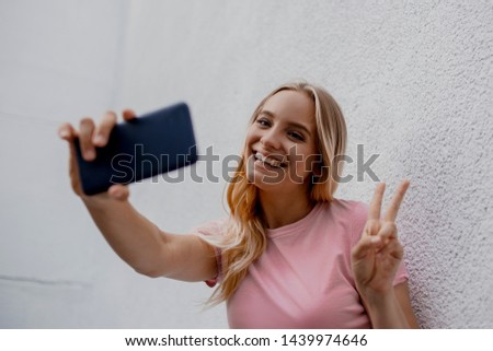 Portrait of attractive teenager girl standing over white street wall, walking, holding smartphone device taking selfies pictures of herself on vacation. People, travel, technology, lifestyle, authenti