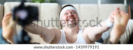 Vlogger freak shows his subscribers on camera how not to do the stretching correctly, holding his hands over his feet. Astonished face expression grimaces aganist home background concept