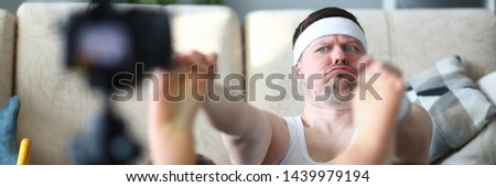 Vlogger freak shows his subscribers on camera how not to do the stretching correctly, holding his hands over his feet. Astonished face expression grimaces aganist home background concept