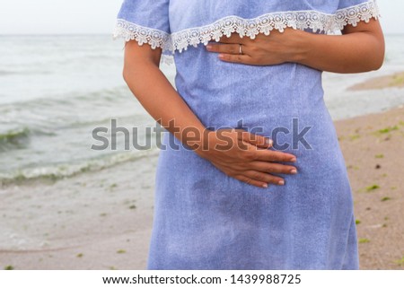 Close up picture of pregnant woman wearing blue dress at the seaside, is holding her tummy 

