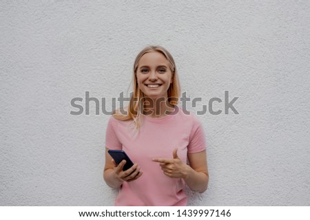 Smiling woman is pointing on smartphone standing on white background looking at the camera with open mouth. People lifestyle. Authentic situation. Picture with copy space.
