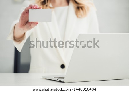 cropped view of businesswoman showing at camera white blank business card while sitting near laptop