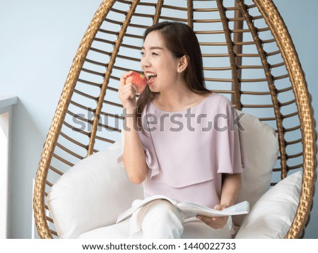 Asian woman eating apple and reading a book while sitting in modern armchair in shape of birds nest at home