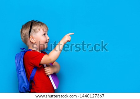Back to school First grade junior lifestyle. Small boy in red t-shirt. Close up studio photo portrait of smiling boy in glasses with schoolbag and book show somethind with his finger.