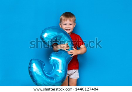 small cute blonde boy on blue background holding foil-coated sphere baloon blue colour. happy birthday three years old.