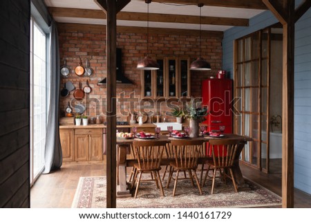 Kitchen in a country house. Wooden kitchen set with big table and chairs standing near the panoramic window to the floor, red refrigerator, blue wooden wall, pans hanging on a brick wall. Loft style. Royalty-Free Stock Photo #1440161729