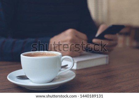Closeup image of casual business man using mobile smart phone connecting free wifi browsing internet with book, latte coffee on wooden table in coffee shop, vintage style.