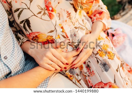 Stylish pregnant couple holding hands on belly in sunny light in summer park. Happy young parents, mom and dad, hugging baby bump and smiling, enjoying beautiful moment at sunset among grass
